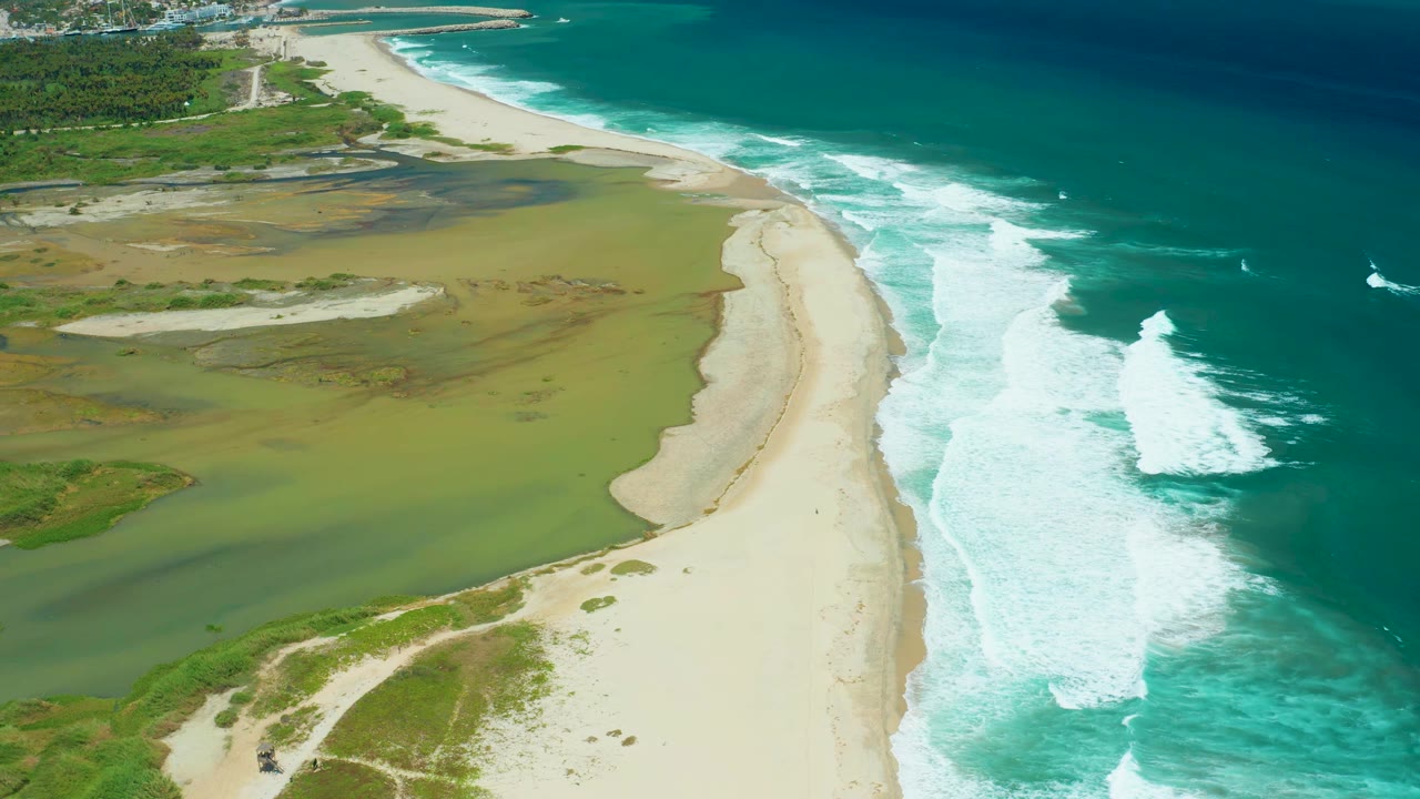San Jose Del Cabo aerial view with foamy ocean waves advancing along the right side of the beach and shallow green wetland water on the left, with no visible people.