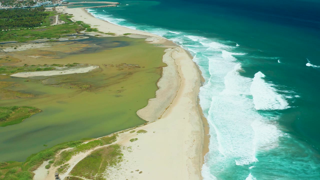 Aerial view over San Jose Del Cabo coastline, where a curved sand strip borders green marsh shallows and bright white breakers on the blue ocean side, with no visible people.