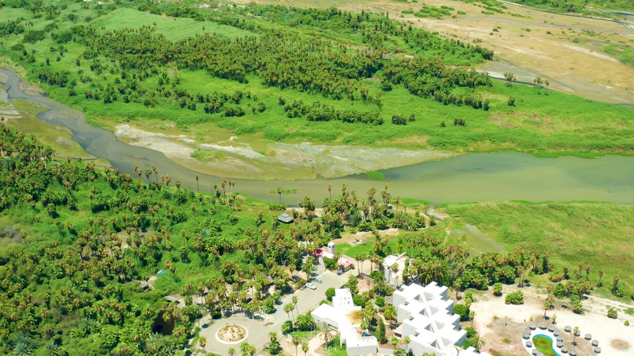 Opening aerial frame in San Jose del Cabo, Baja California Sur, showing a white angular-roofed building at the lower edge, palms and dense greenery around it, a pale river channel across the middle, and no visible people.