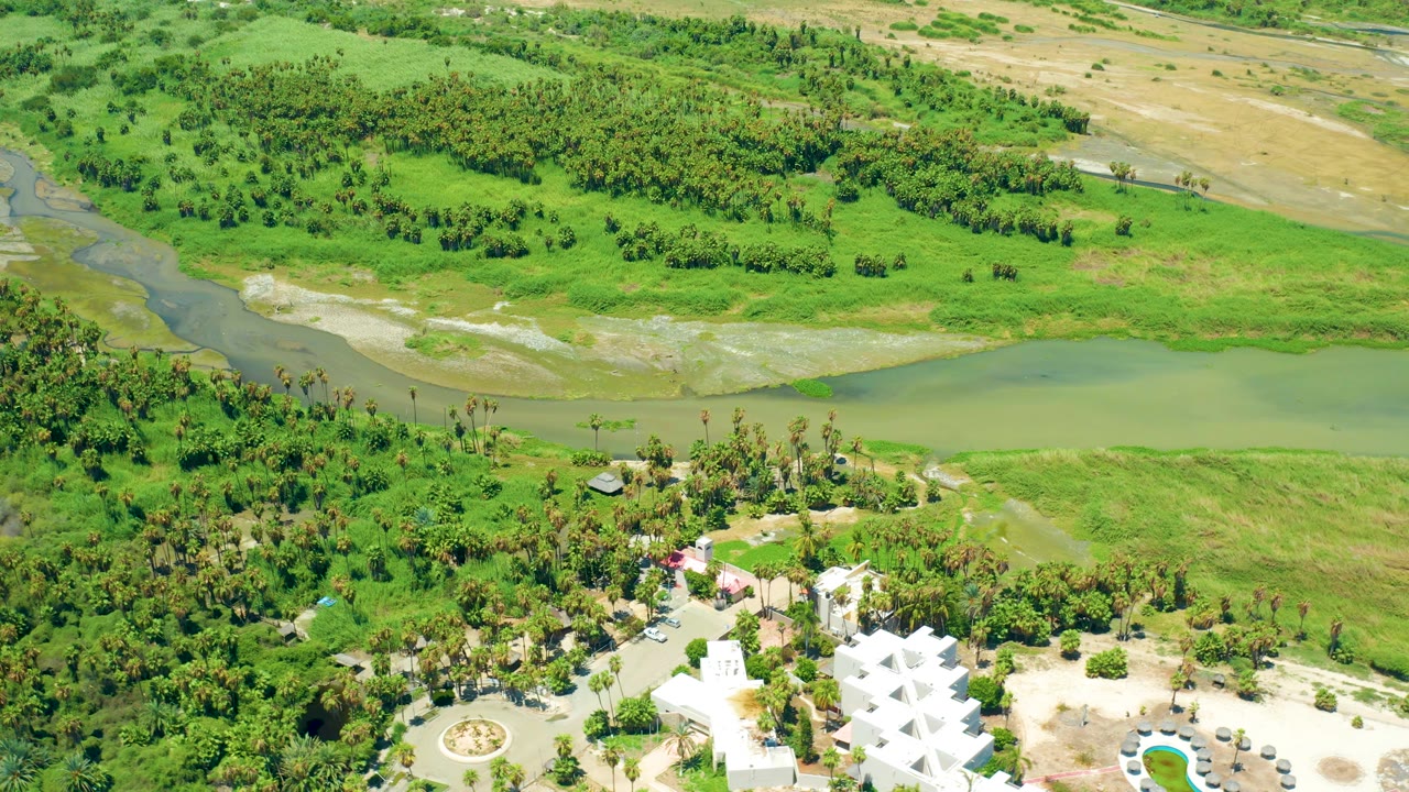 Third frame shows San Jose del Cabo greenery from above, with the white geometric building partly cropped at the bottom, a tree band behind it, a winding river channel, and no visible people.