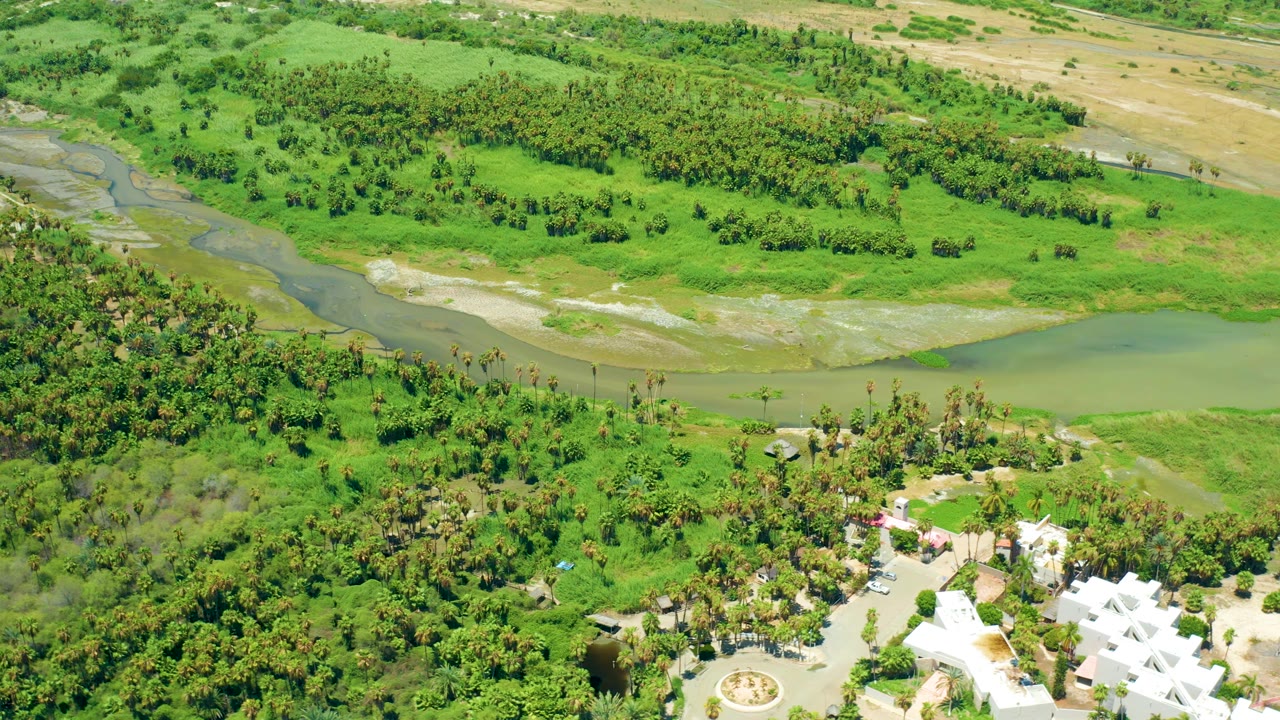 This San Jose del Cabo aerial frame keeps the river and pale sandbars across the upper area while white resort buildings and planted grounds sit along the lower right, with no visible people.