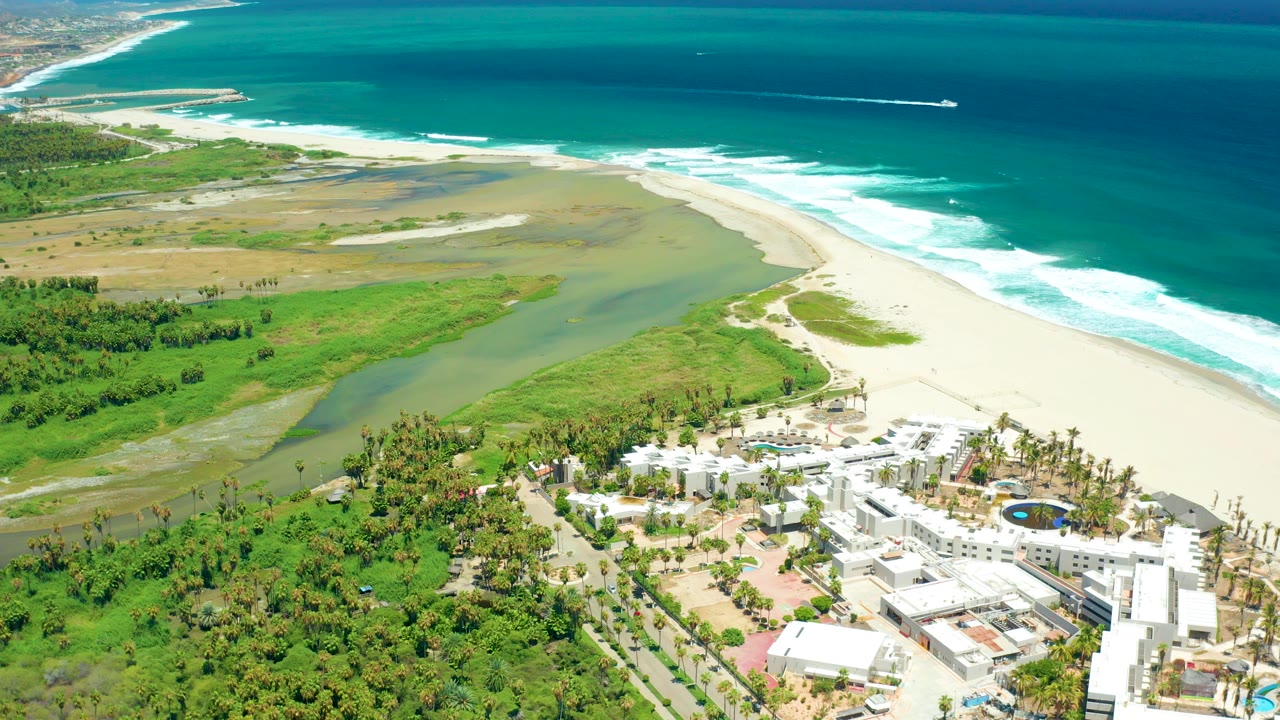 Aerial view over San Jose del Cabo showing a green lagoon beside pale sand, white surf, turquoise ocean, resort buildings, and a small offshore boat, with no visible people.