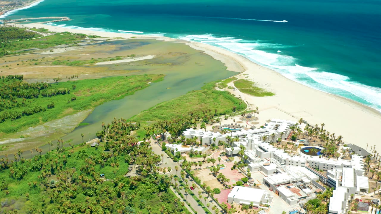 Aerial frame over San Jose del Cabo with the lagoon widening across green terrain, white sand meeting foamy surf, and resort buildings at the bottom edge, with no visible people.