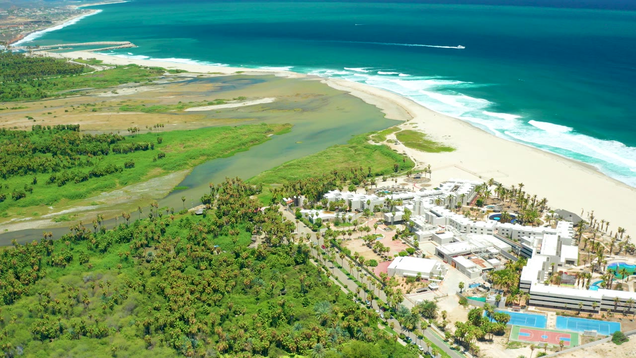 Aerial view over San Jose Del Cabo with green wetland, pale beach, turquoise ocean surf, white shorefront buildings, pools, and no visible people.