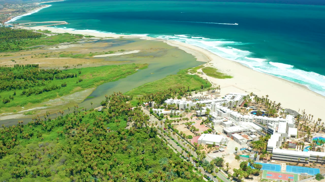 Aerial view in San Jose Del Cabo showing the wetland flats and beach curve, with white buildings and pools at the lower right edge and no visible people.