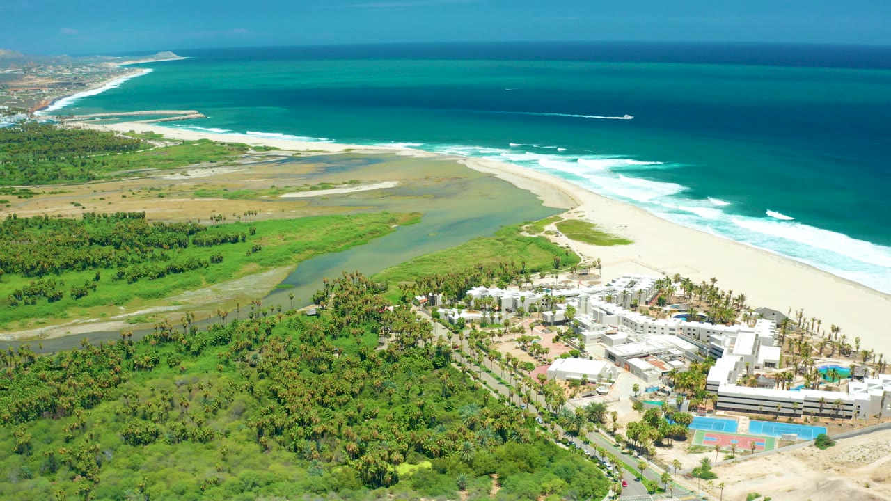 High aerial frame over San Jose Del Cabo showing green coastal land, a river mouth, pale beach, turquoise ocean, white surf, resort buildings, and a small offshore boat wake with no visible people.