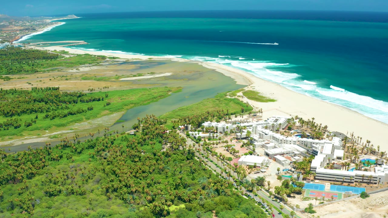 Aerial coastline with river mouth, green lowland, buildings, beach, surf, and offshore wake