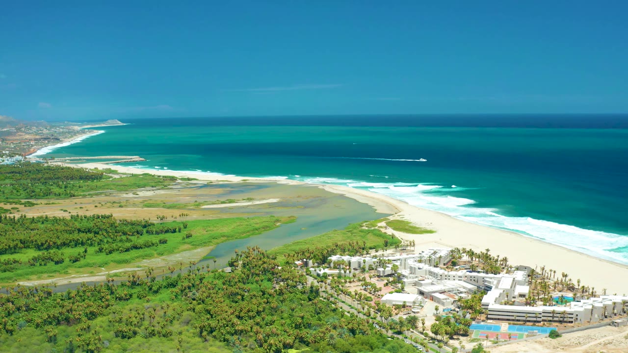 Wide San Jose del Cabo aerial showing the river mouth meeting the beach, resort buildings below, a small white boat offshore, and no visible people.