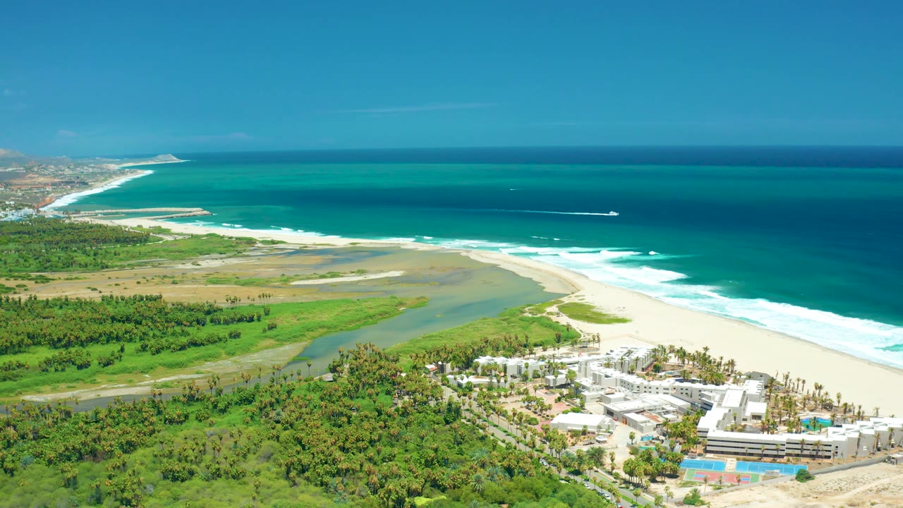San Jose del Cabo aerial frame with the surf line bright along the beach, the river channel behind it, green coastal vegetation, resort buildings, and no visible people.