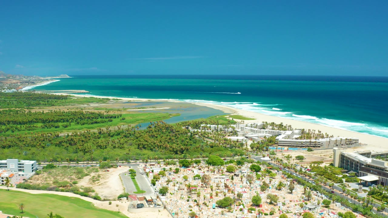 Aerial frame over San Jose del Cabo, Mexico showing turquoise ocean, white surf, sandy beach, green vegetation, coastal buildings, parking areas, and a small offshore boat with no visible people.