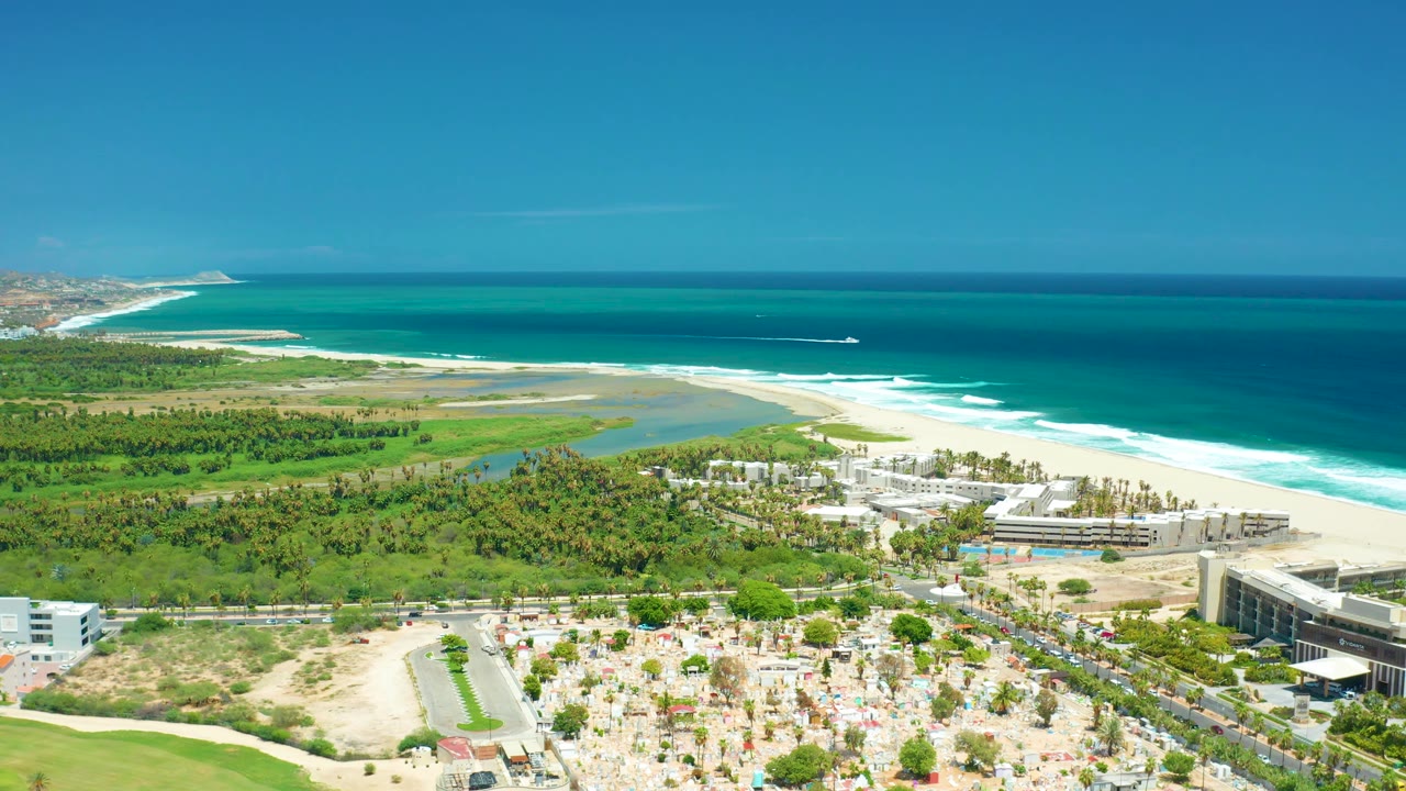San Jose del Cabo coastline appears from above with layered blue ocean, a white surf break, sandy beach, green vegetation, beachfront buildings, parked cars, and no visible people.