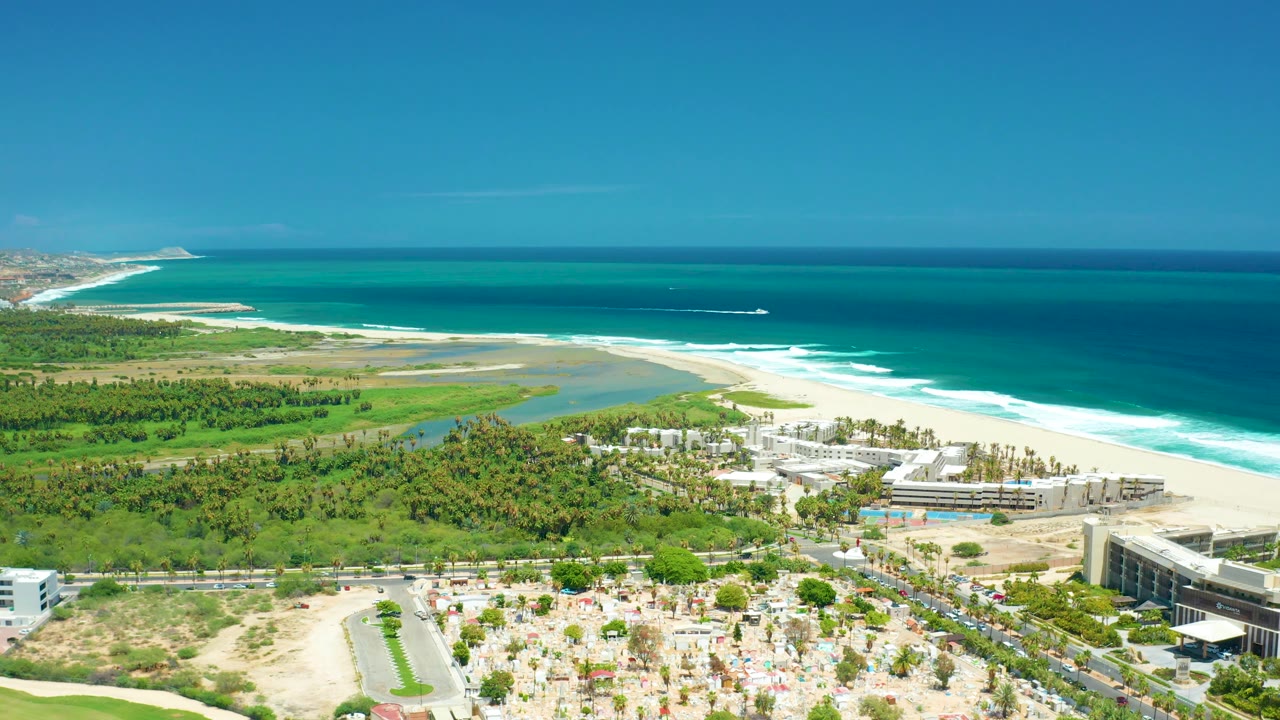 A wide aerial frame over San Jose del Cabo shows more open sky above turquoise ocean, beach surf, green coastal land, white buildings, roads, parked cars, and no visible people.