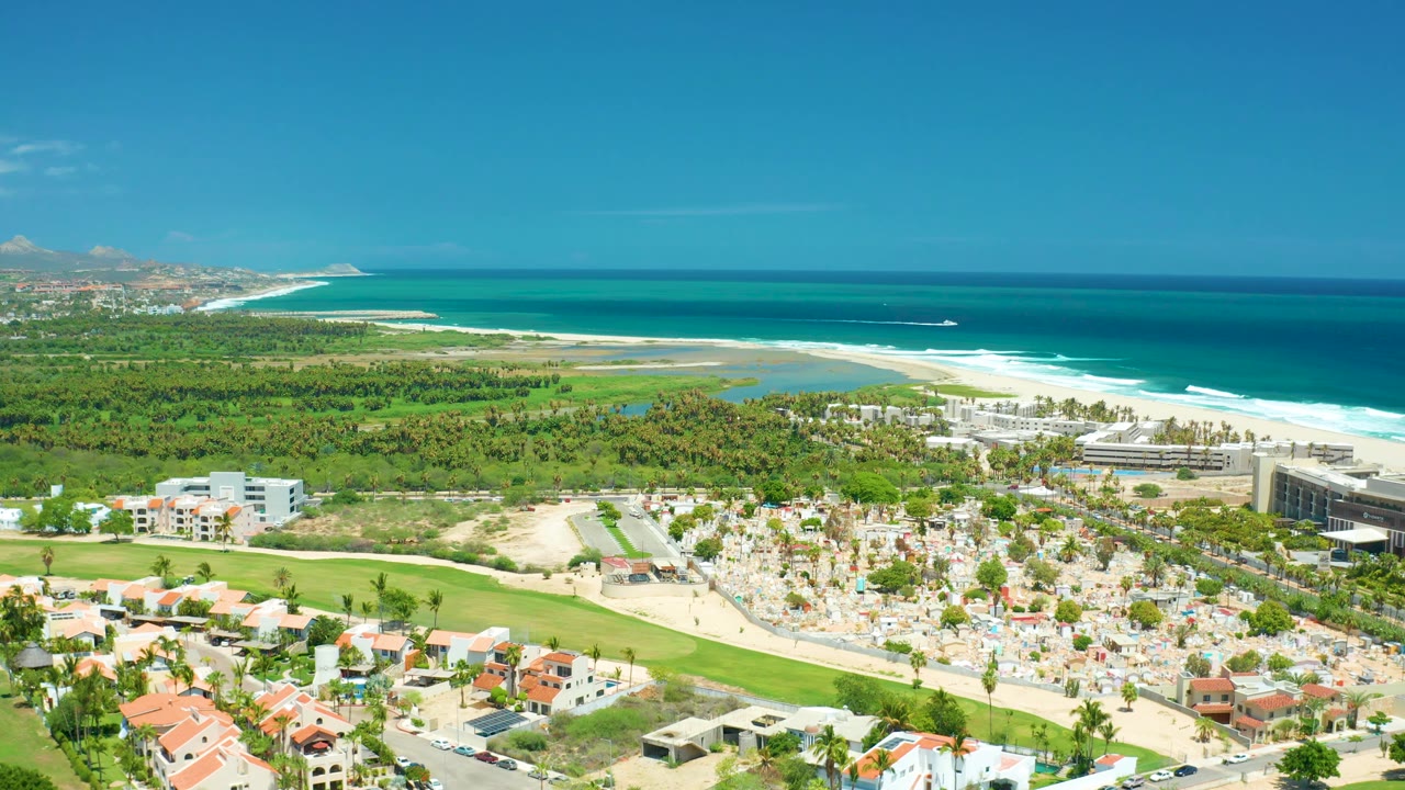 San Jose del Cabo aerial view with more coastline visible to the left, turquoise water and surf beyond the sand, resort buildings on the right, and small figures among umbrellas below.