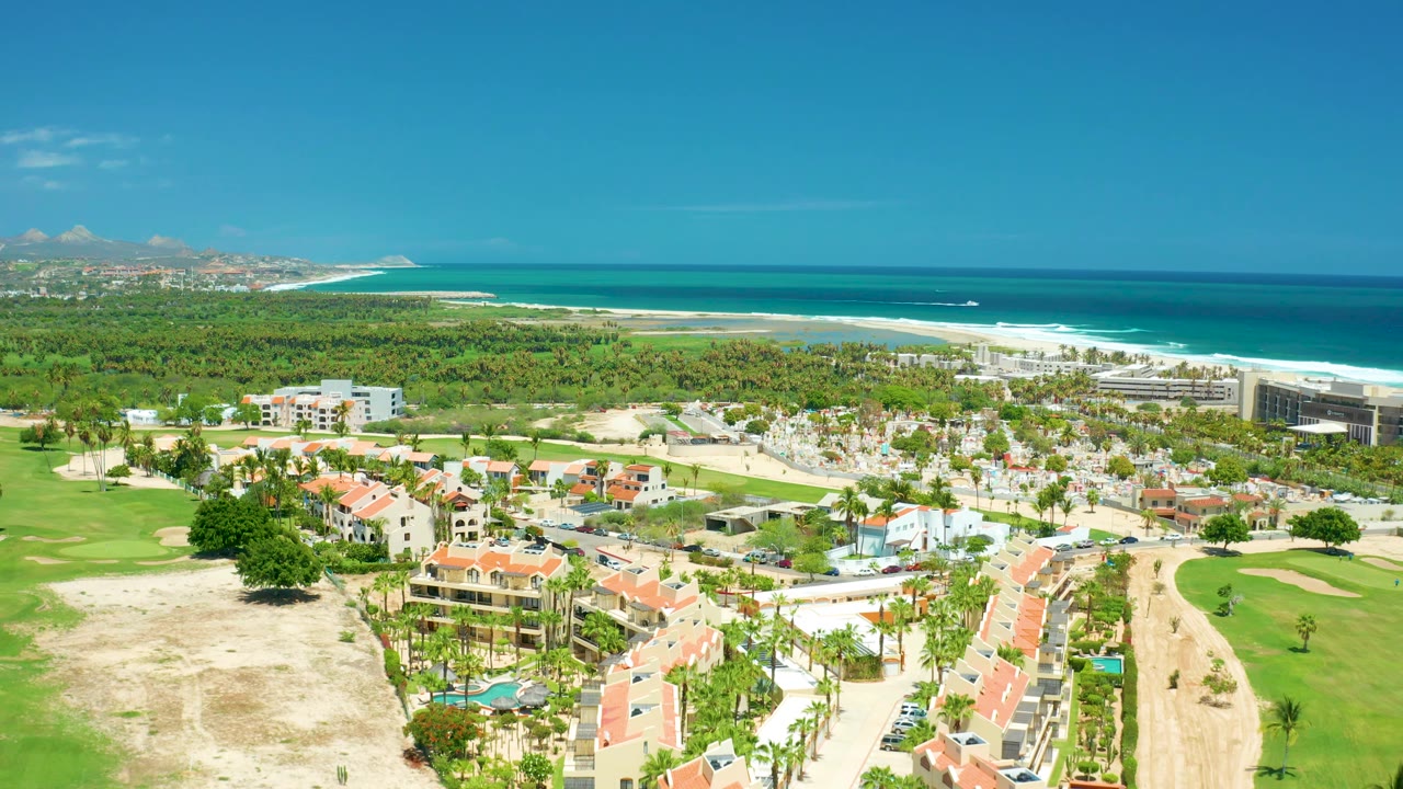 Later aerial frame in San Jose Del Cabo with a broad green foreground, clustered red roofs, beachfront development to the right, surf lines along the beach, blue ocean, and no visible people.