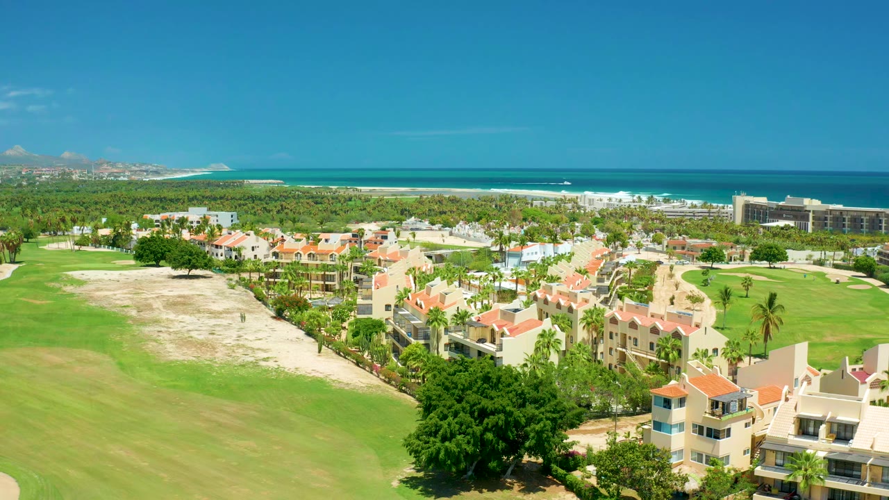 Wide aerial over golf greens, resort buildings, palm trees, beach, and ocean.