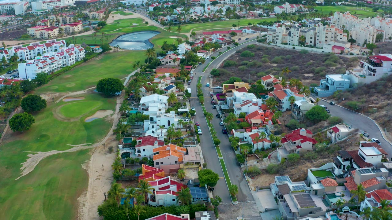 Opening aerial frame over San Jose del Cabo shows golf fairways and sand traps beside red-roofed and white homes, a curving residential road, dry scrub lots, and no visible people.
