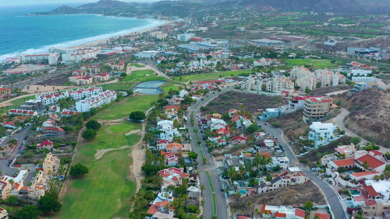 Aerial frame over San Jose del Cabo showing a broad green golf course at lower left, clustered residential buildings, curving streets, the shoreline to the upper left, and no visible people.