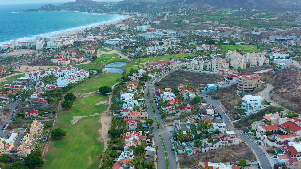 San Jose del Cabo aerial view with white residential blocks near the center, green golf fairways below, a pale beach and turquoise ocean along the left edge, hills in back, and no visible people.