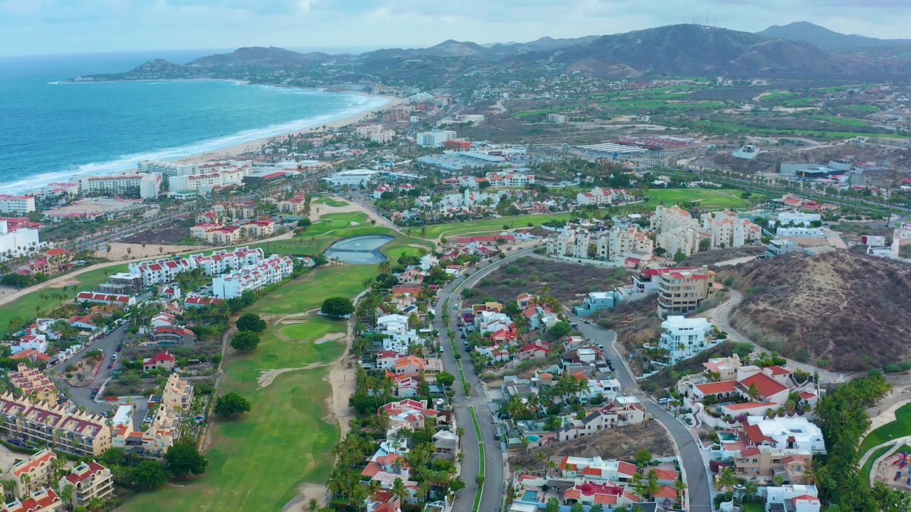 Wide aerial frame in San Jose del Cabo showing golf course greens and ponds below, compact residential blocks across the center, the ocean shoreline at left, and no visible people.