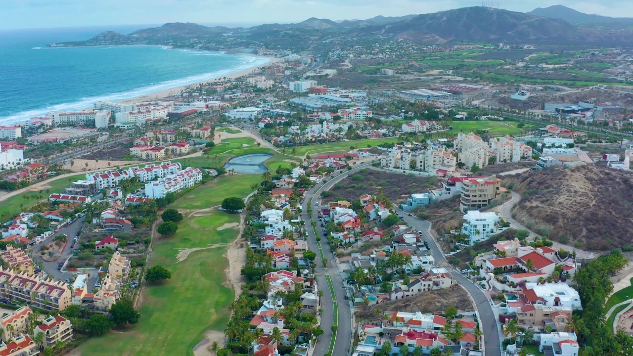 San Jose del Cabo aerial view with fairways and roads lower in frame, dense homes and low-rise buildings through the middle, beach and blue ocean to the left, and no visible people.