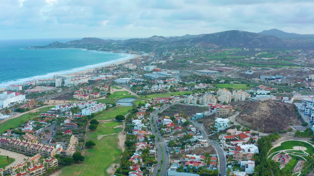 Wide aerial view over San Jose Del Cabo with turquoise ocean and surf at left, beachside buildings, golf course greens, residential streets, dry hills, distant mountains, cloudy sky, and no visible people.