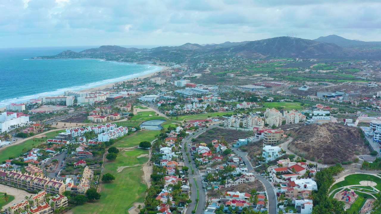 Aerial view in San Jose Del Cabo with bright ocean water at left, a pale sand beach, low-rise buildings, residential streets, green fairways and ponds in the lower frame, mountain ridges in back, and no visible people.