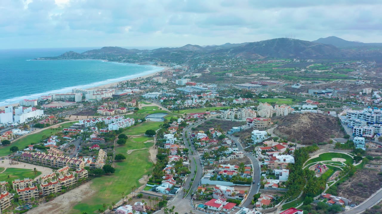 Aerial coastal landscape with ocean, beach, green fairways, roads, homes, and distant hills.