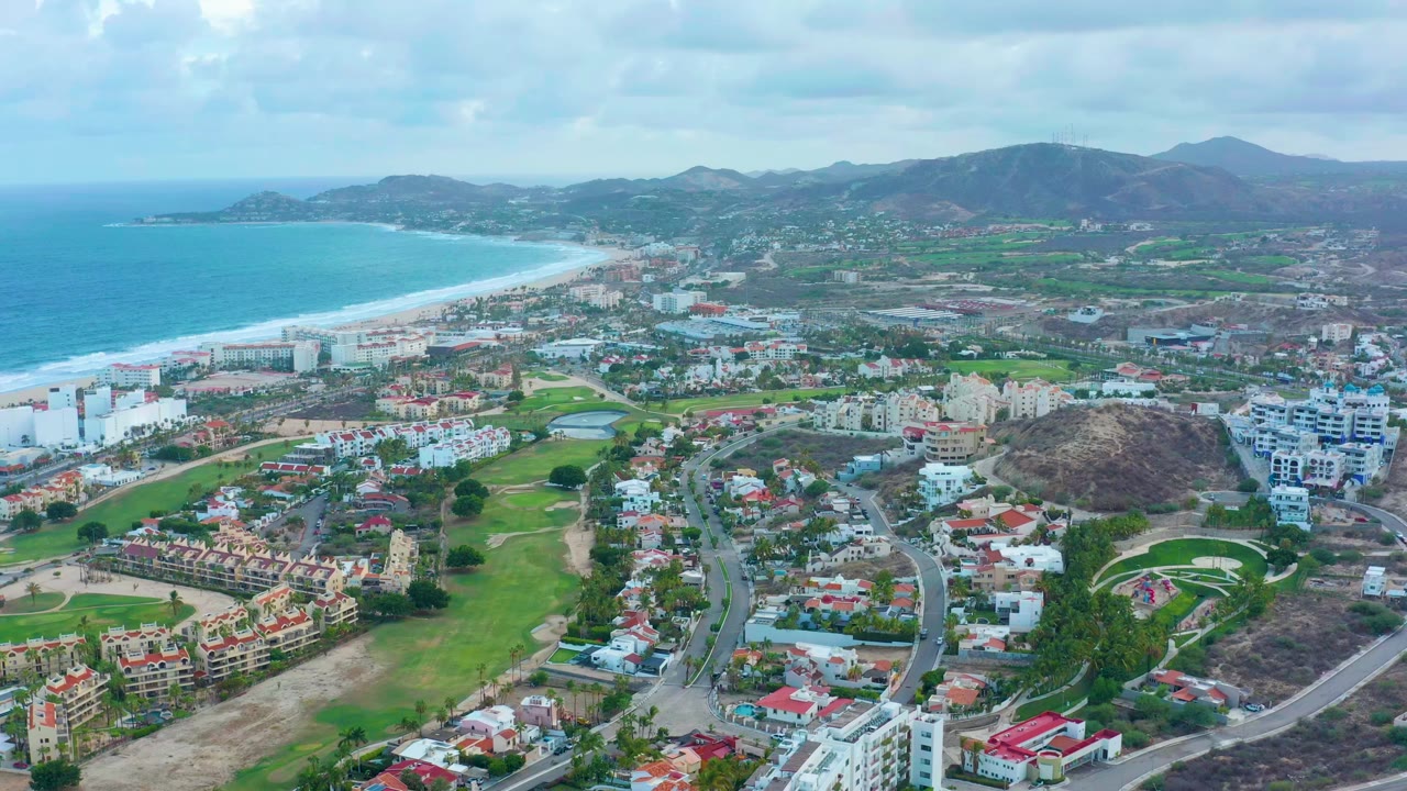 Opening aerial frame over San Jose Del Cabo with turquoise ocean and sandy beach at left, town blocks and green spaces across the center, hills and mountains beyond, and no visible people.