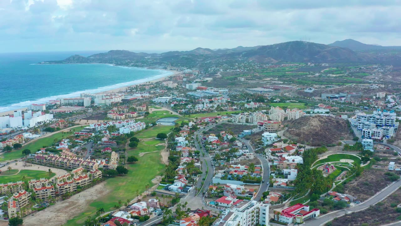 Wide drone view of beach, ocean, roads, homes, green spaces, hills, and cloudy sky.