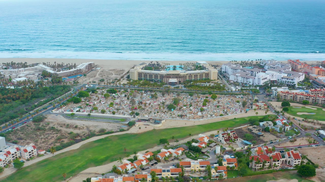 Opening aerial frame over San Jose Del Cabo, Mexico, with blue ocean beyond a sandy beach, resort buildings at midframe, green land and red-roofed houses below, and no visible people.