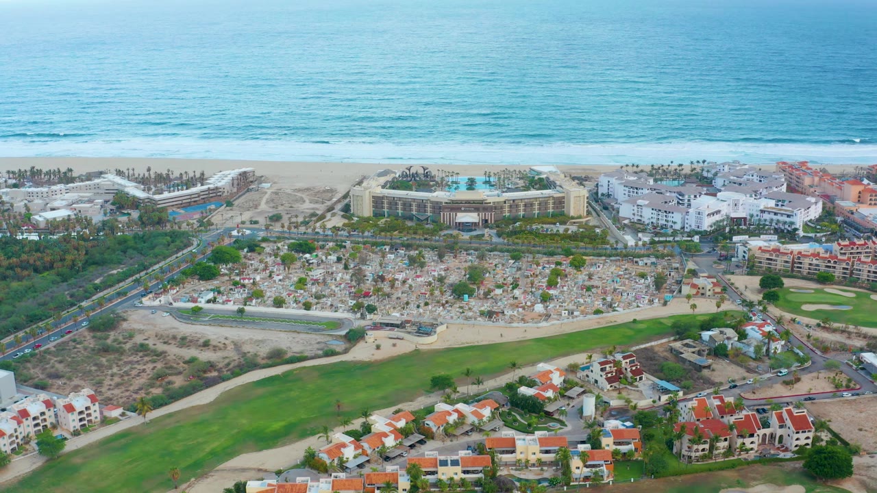 Wide aerial over beachside resort buildings, ocean, roads, green land, and homes.