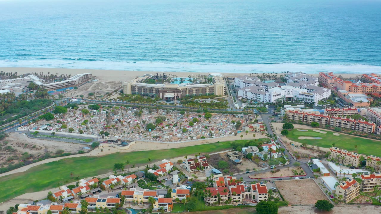 Aerial frame in San Jose Del Cabo showing the ocean horizon, beach strip, large coastal buildings, green golf course, roads, red-roofed residences, and no visible people.