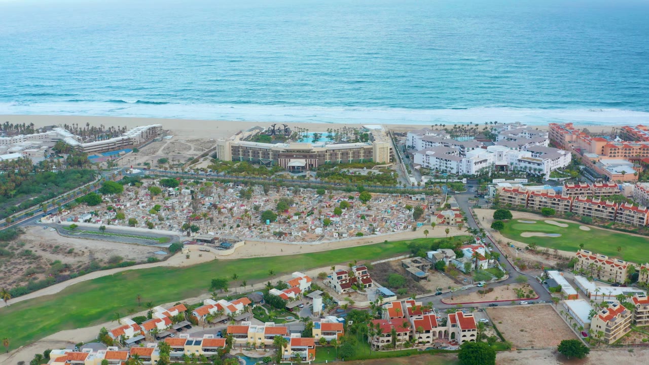 San Jose Del Cabo aerial view with blue ocean above a sandy beach, resort-style buildings, a central demolition area, golf fairway, red-roofed homes, and no visible people.