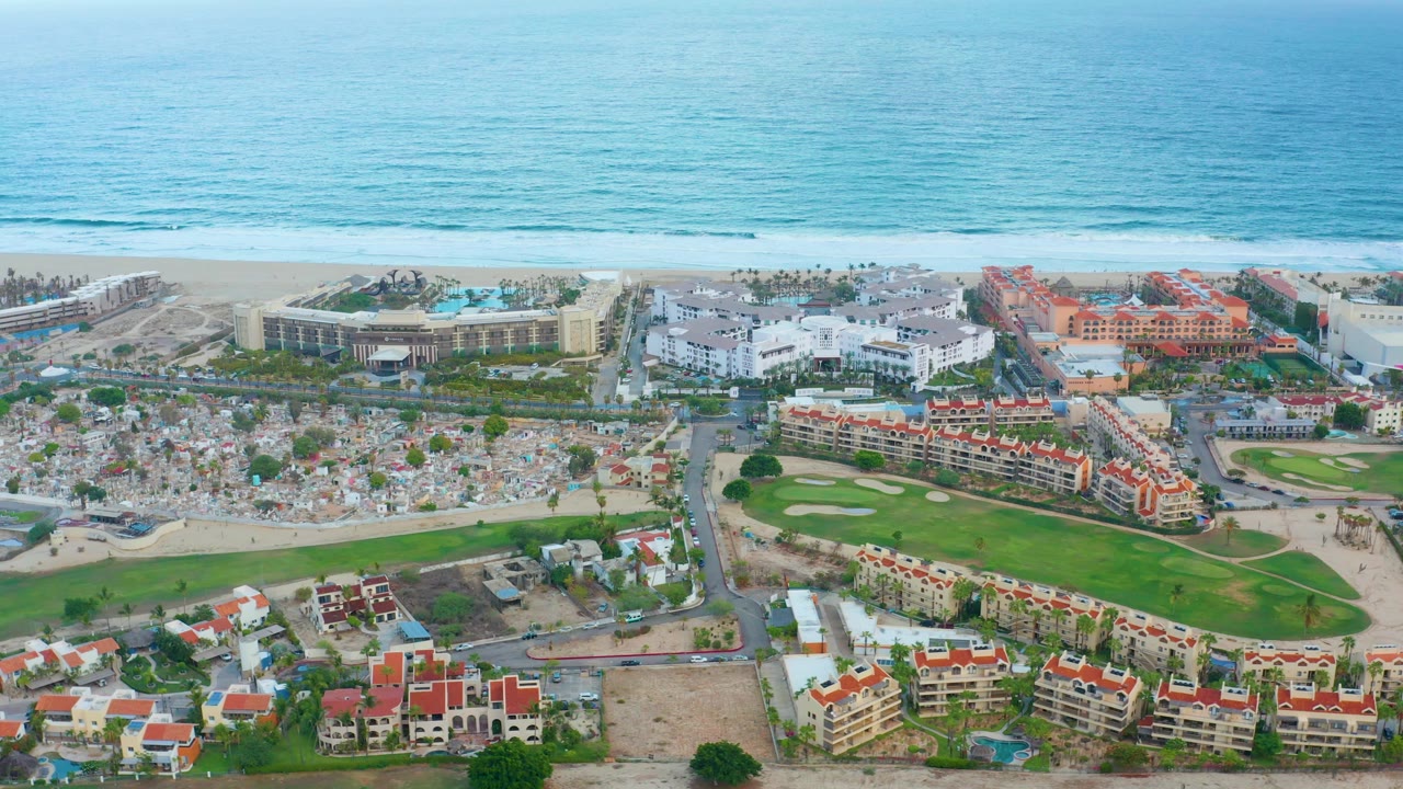 High aerial view over San Jose Del Cabo with blue ocean, sandy beach, beach umbrellas, resort buildings, a road, and a green golf course in the foreground.