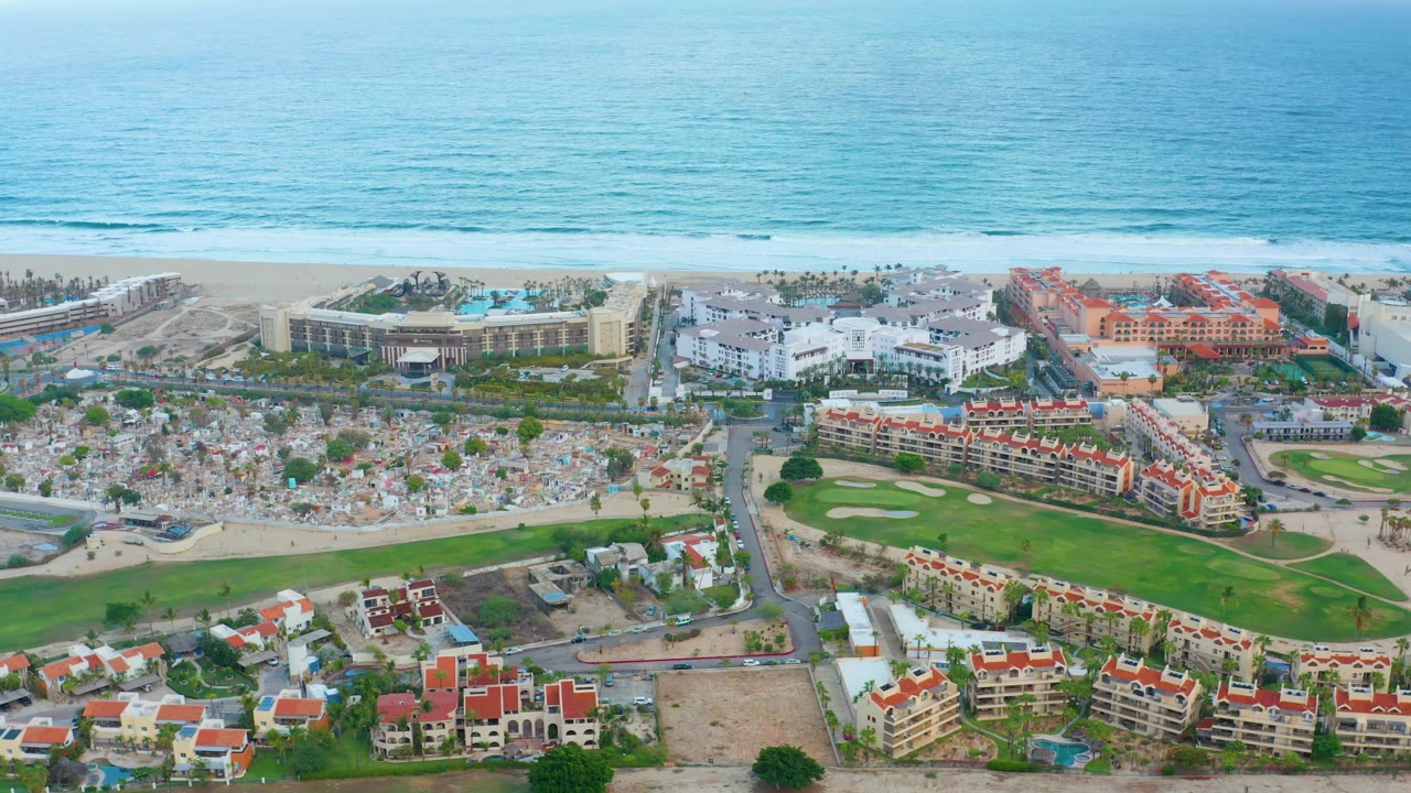 Aerial view in San Jose Del Cabo showing a bright blue ocean horizon, crowded beach umbrella area, white resort buildings, orange-red roofs, and a green golf course below.