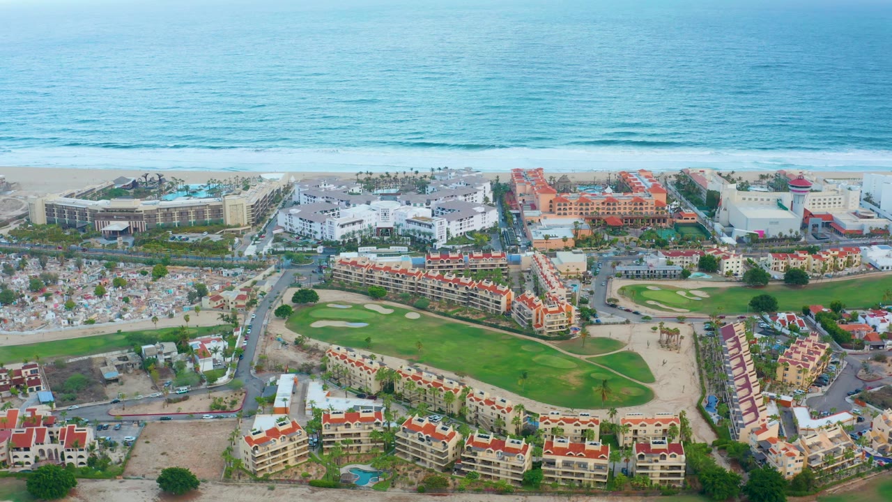 The second frame keeps the ocean high in the view while San Jose del Cabo resort blocks, parking areas, red-roofed buildings, and golf fairways fill the lower half with no visible people.