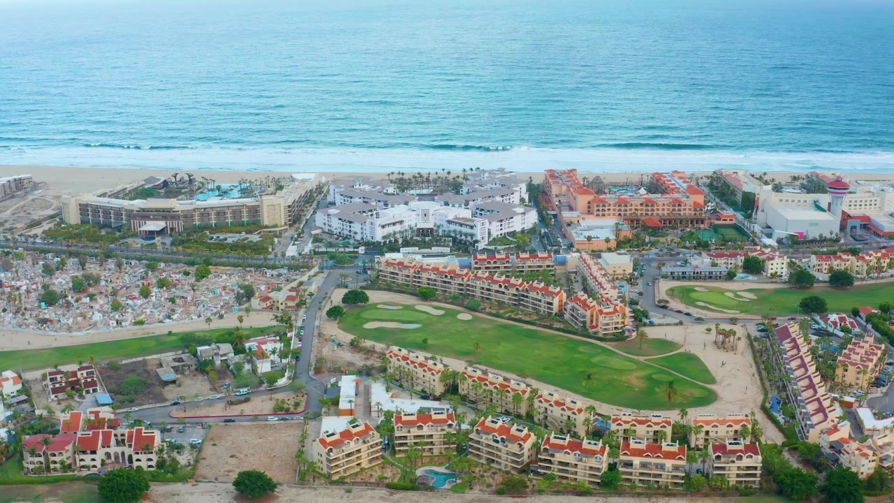 Mid-clip, the San Jose del Cabo coastline remains centered with blue water, pale beach, multi-story resort buildings, parking areas, roads, and a green golf course with no visible people.