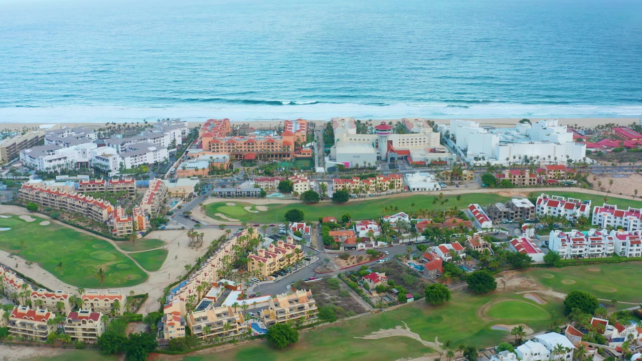 Opening aerial frame of San Jose Del Cabo coastal residences, green golf course areas, sandy beach, and blue ocean in the background, with no visible people.