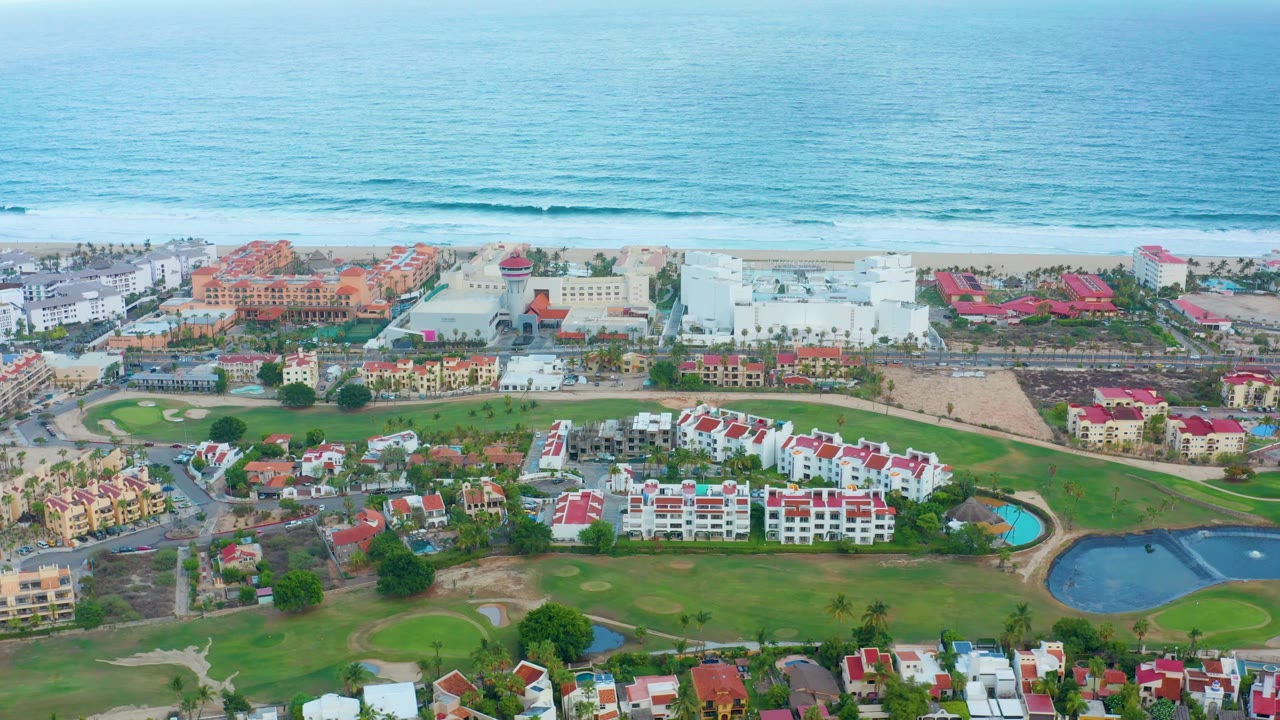 Wide aerial frame in San Jose Del Cabo showing red-roofed residential buildings, green grounds, larger beachfront structures, a sandy shoreline, blue ocean bands, and no visible people.