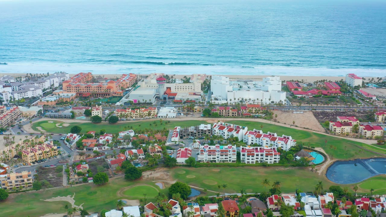 San Jose Del Cabo aerial still with white and red-roofed buildings around green lawns, a curved blue pool or pond at right, sandy beach, ocean waves, and no visible people.