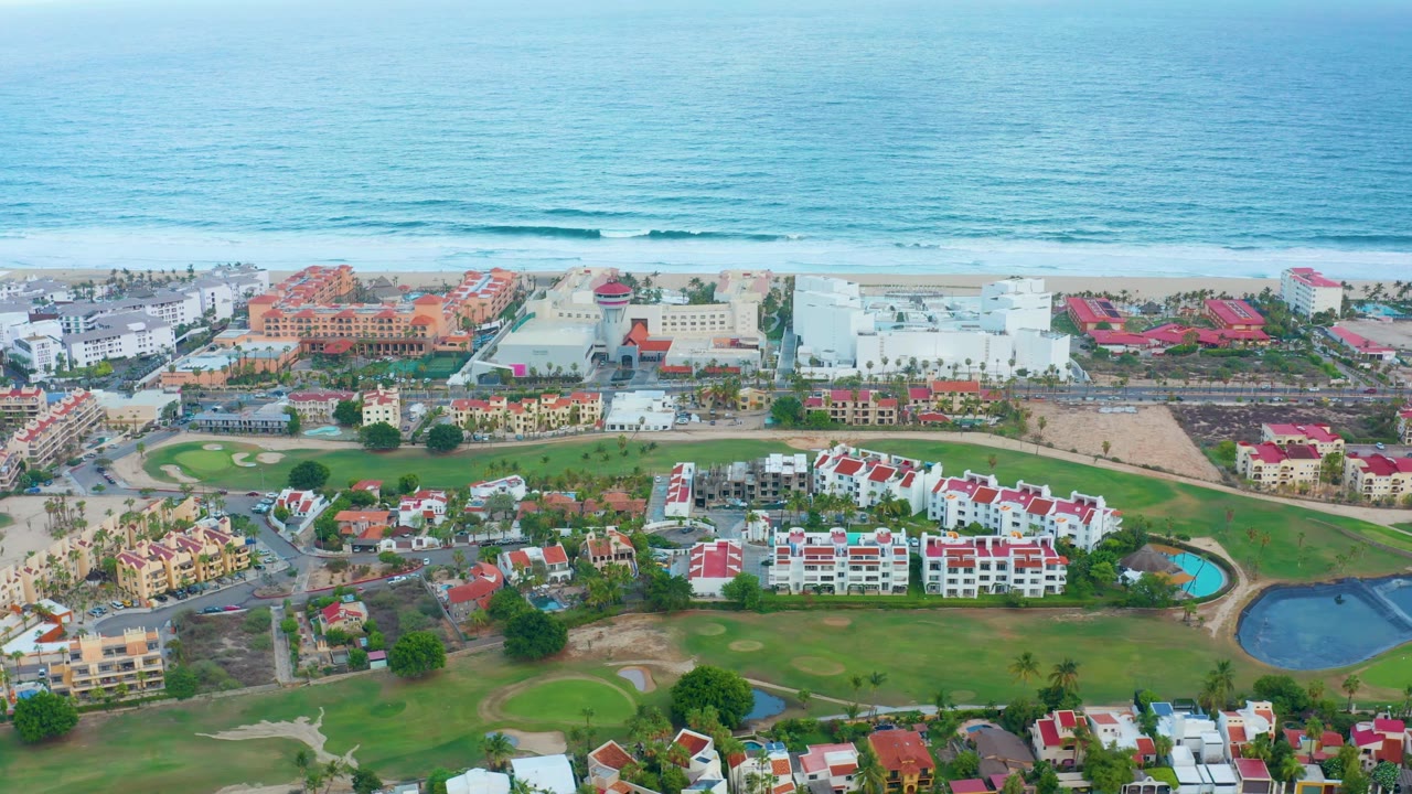 Coastal buildings and green grounds below a sandy beach and calm ocean.