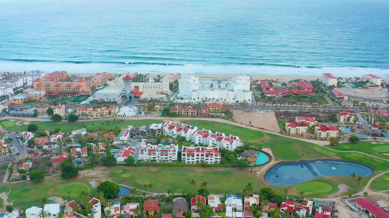 Wide aerial frame over San Jose del Cabo showing blue ocean and surf behind a beach, resort and residential buildings, green golf fairways, two ponds, roads, palms, and no visible people.
