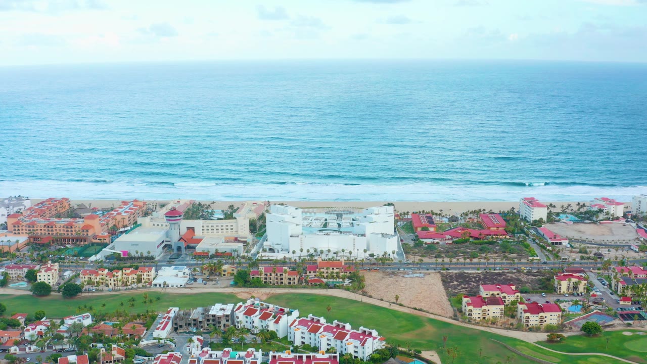 Wide aerial view of San Jose del Cabo resort buildings, green foreground grass, sandy beach, blue ocean, light clouds, and no visible people at the opening moment.