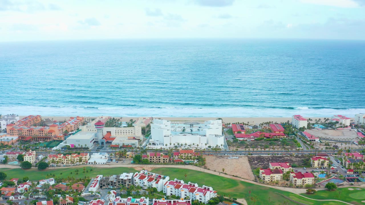 San Jose del Cabo aerial view with resort buildings and red rooftops across the lower frame, a pale sandy shoreline, broad blue ocean, and no visible people.