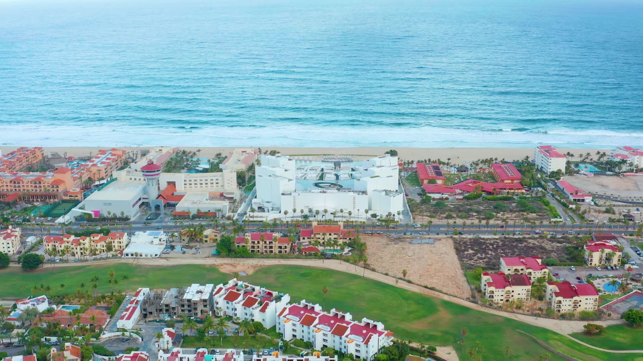 San Jose del Cabo aerial frame with the ocean filling the upper half, pale surf along the beach, red-roofed resort buildings and green lawn areas below, and no visible people.