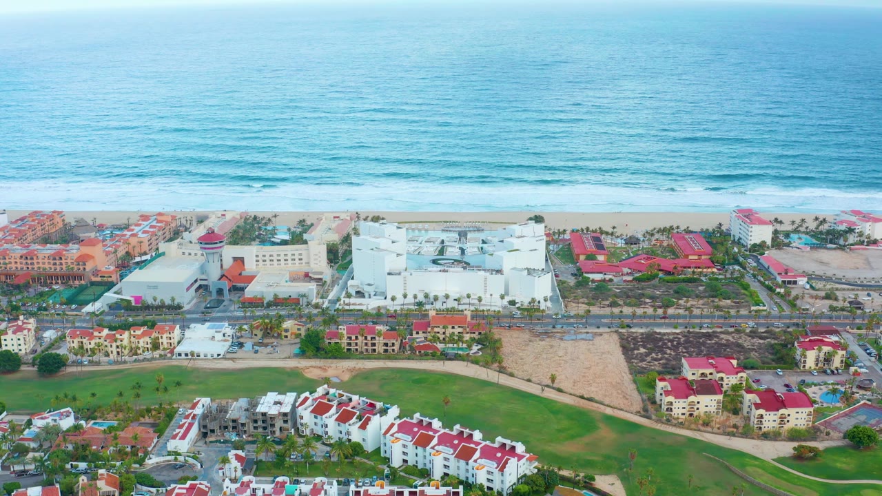 Aerial view over San Jose del Cabo resort grounds, with white buildings and red roofs in front of a sandy beach, rolling ocean waves, hazy sky, and no visible people.