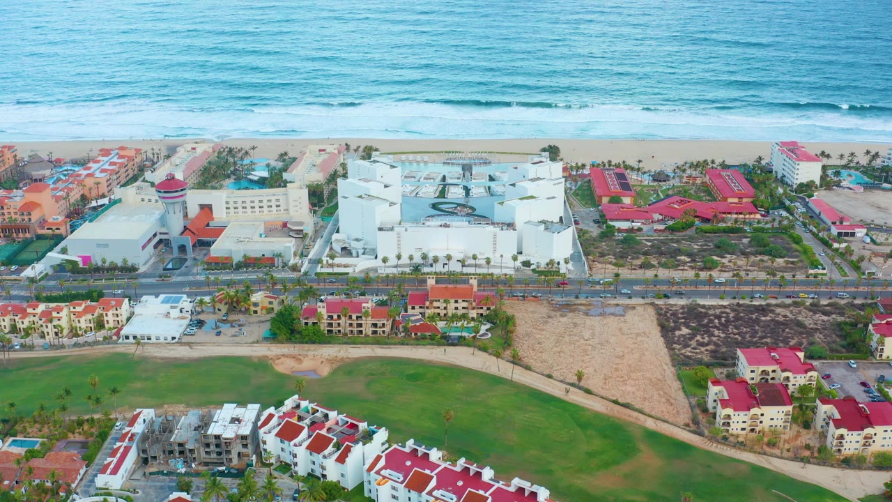 At 0:00, an elevated San Jose Del Cabo view shows blue ocean, a thin sandy beach, white resort-scale buildings, red rooftops, a road, green fields, and no visible people.