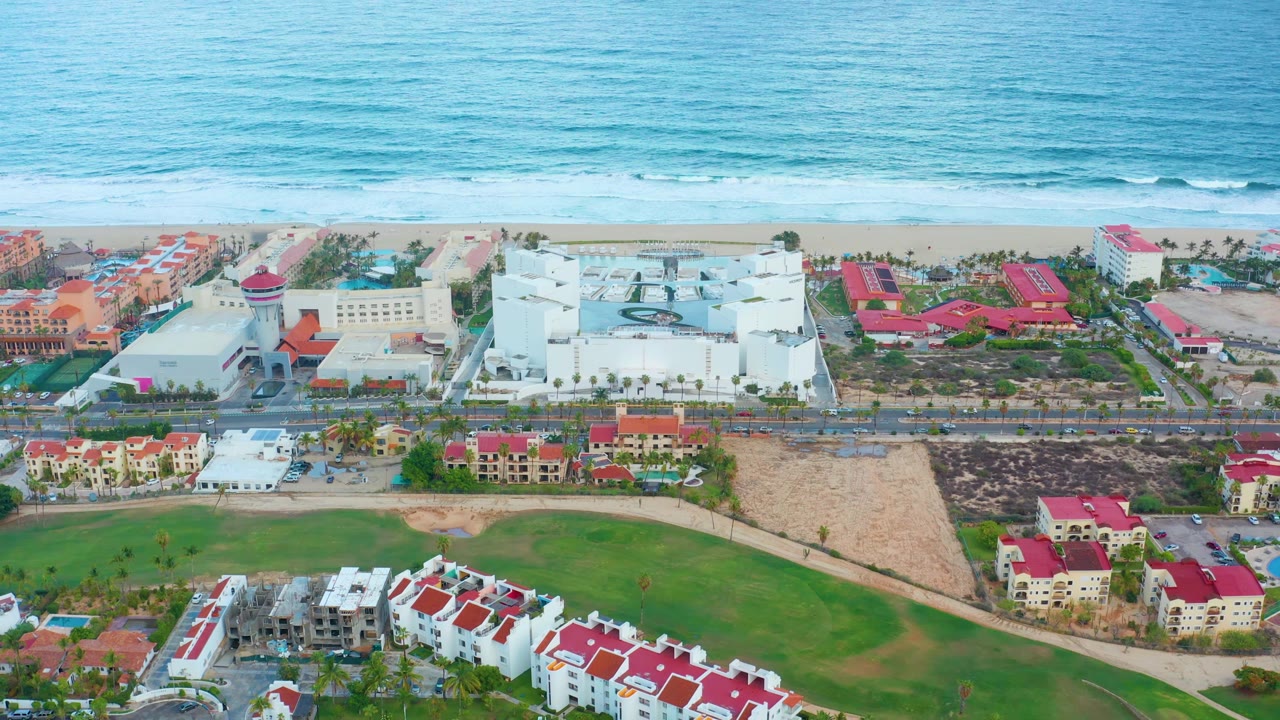 Elevated view of beach, surf, white coastal buildings, red roofs, road, and green space.