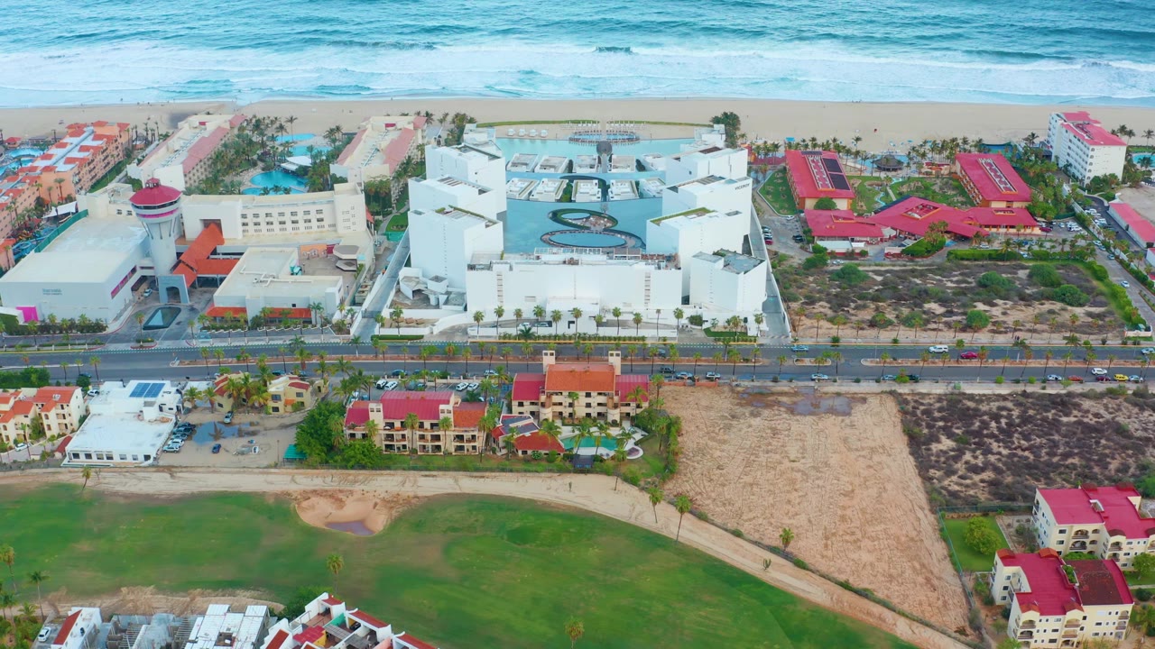 At 0:01, the San Jose Del Cabo aerial frame holds the ocean and beach above a white resort, visible pool area, roadside traffic, red roofs, lawns, and no visible people.