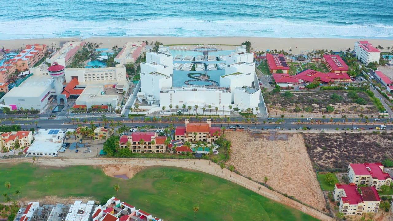 At 0:02, San Jose Del Cabo appears from above with the surf line, sand, white resort buildings, a central pool area, road traffic, green lawns, and no visible people.